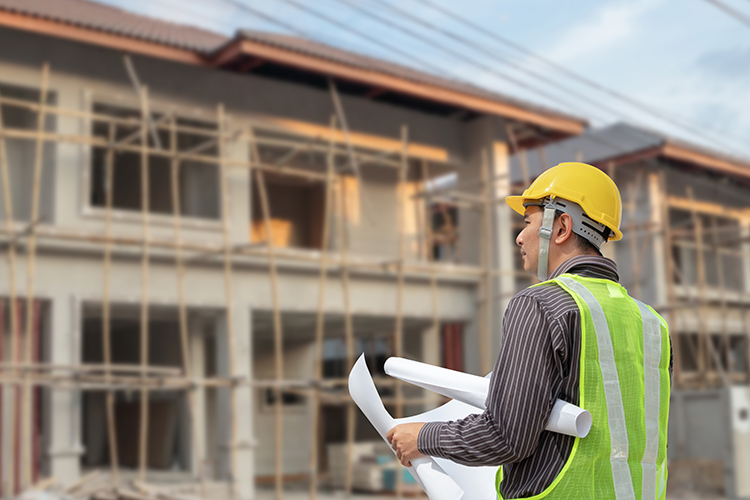 Man in construction hat and safety vest holding blueprints looking up at a home under construction
